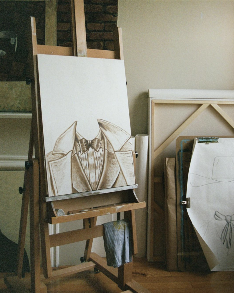 Easel featuring a detailed sketch of a suit jacket and shirt, with additional art materials and canvases leaning against a wall in a studio setting